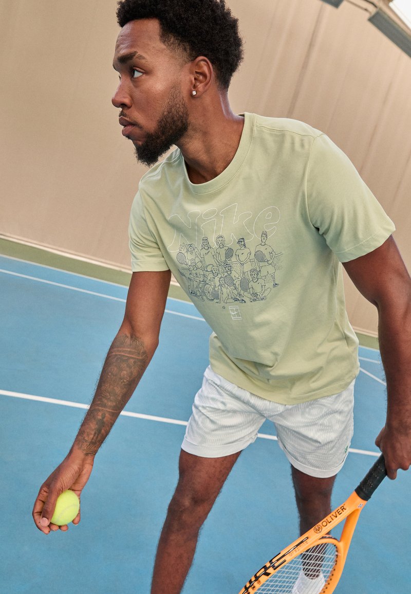 Nike heles roheline t-särk, millel on graafiline trükk, kantud valgete mustriliste lühikeste pükste juurde. Hoiab kollast tennisepalli; tennisereket on nähtav.