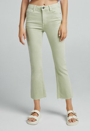 Jasnozielone, krótkie dzwony z surowymi wykończeniami, noszone z białymi sandałami na zatrzask, na stojącej osobie.