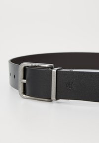 Cintura in pelle nera con fibbia metallica argento, dotata di incisione del logo centrale e multiple aperture per la regolazione. Texture liscia e design resistente.