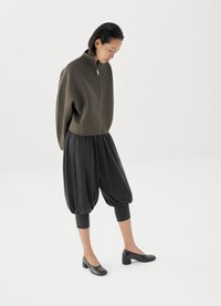 Sweat-shirt zippé vert olive avec pantalon noir style ballon. Chaussures à talons noires avec motif crocodile. Textures lisses et coupe décontractée.