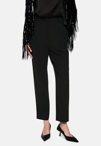 Schwarze, maßgeschneiderte Hose mit Falten und glatter Textur. Kombiniert mit einer schwarzen, mit Pailletten besetzten Fransenjacke und funkelnden schwarzen Absätzen.