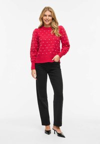 Femme souriante aux longs cheveux blonds portant un pull rouge texturé, un pantalon noir droit et des mules à talons noires, debout devant un fond blanc.