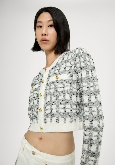 Cardigan court blanc et gris avec un motif à carreaux texturé, boutons dorés et encolure ronde. Présente une coupe ajustée et un tissu tricoté.