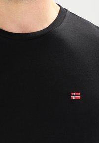 Czarna koszulka z okrągłym dekoltem, z małym haftowanym emblematem norweskiej flagi w kolorach czerwonym, białym i niebieskim na lewej stronie klatki piersiowej.