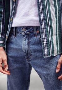 Blue denim jeans with a high waist, featuring contrasting orange stitching, metal button closure, and a textured finish. Striped green shirt detail visible.