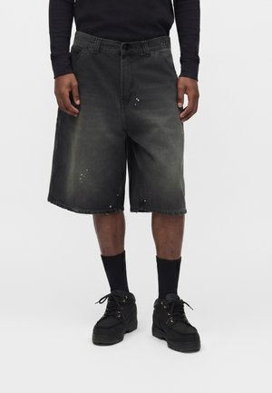 Man draagt vervaagde zwarte denim shorts tot op de knieën, zwart shirt met lange mouwen, zwarte sokken en zwarte veterschoenen, staand tegen een witte achtergrond.