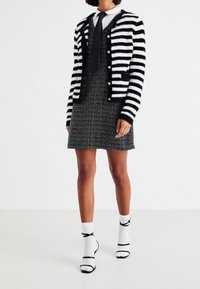 Svartvit randig kofta med guldfärgade knappar över en svart klänning med textur. Kombinerad med vita sockor och svarta skor med rem.