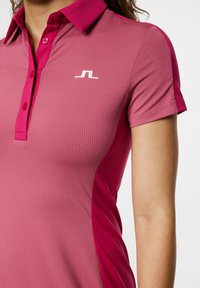 Pikétröja i ljusrosa med texturerad finish. Har en kontrasterande mörkrosa axeldetalj och en vit logotyp på vänster bröst.