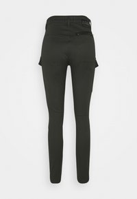 Musta slim-fit housut korkealla vyötäröllä, vetoketjullinen tasku oikealla takana ja sivussa cargo-taskut, esitetty takaa valkoista taustaa vasten.