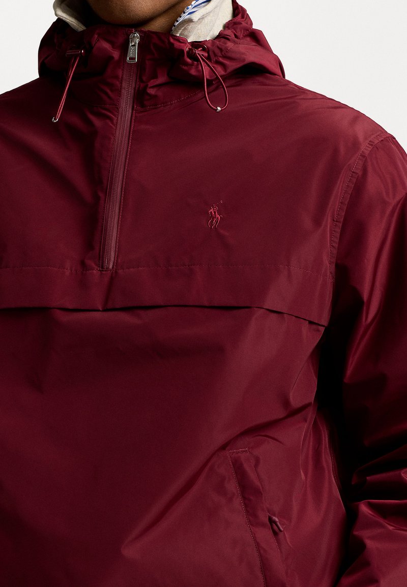 Burgunderfarbene Windjacke mit halbem Reißverschluss, Kapuze mit Kordelzügen und einer Fronttasche. Hergestellt aus leichtem, glattem Stoff mit Logodetail.