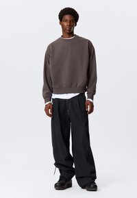 Dunkelgrauer Pullover-Sweatshirt aus weichem Stoff, mit Rundhalsausschnitt und gerippten Bündchen. Getragen über einem weißen T-Shirt und kombiniert mit dunklen Hosen.