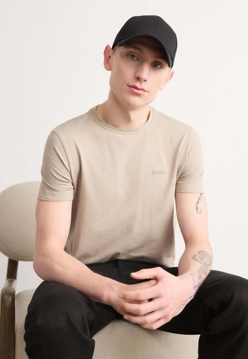 Μπεζ κοντομάνικο μπλουζάκι με στρογγυλή λαιμόκοψη, με μικρή ραμμένη λεπτομέρεια. Συνδυασμένο με μαύρο παντελόνι και μαύρο καπέλο. Απλός, καθημερινός σχεδιασμός.