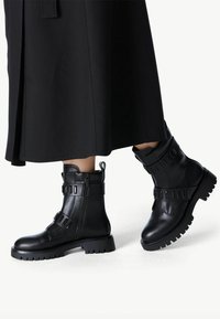 Black leather combat boots with a rubber sole, featuring a side zipper, strap accents, and a textured upper with a sturdy design.