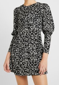 Svart klänning med vitt blommönster, långa puffärmar, rund halsringning, åtsittande midja och utsvängd kjol. Slätt tyg med lätt lyster.