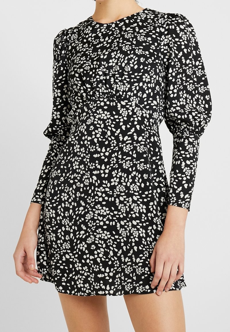 Svart klänning med vitt blommönster, långa puffärmar, rund halsringning, åtsittande midja och utsvängd kjol. Slätt tyg med lätt lyster.