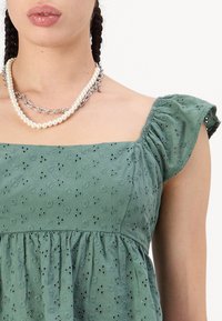 Vestido de algodón verde con mangas cortas abullonadas, escote cuadrado y patrón de ojetes florales. Collar de perlas en capas y cadena de metal.
