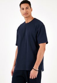 Tricou de culoare bleu marin, cu mânecă scurtă, fabricat dintr-un material moale, având guler rotund și o croială relaxată cu mâneci răsucite.