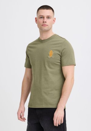 Ung mand med kort hår iført en olivengrøn T-shirt med en lille orange tegneseriefigur på venstre bryst, kombineret med sorte bukser.