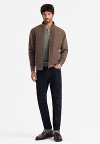Bruine gewatteerde bomberjack over een lichtgroen henley-shirt, gecombineerd met donkerblauwe jeans en bruine bootschoenen. Zachte stof, casual fit.