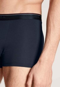 Marineblauwe boxershorts van rekbaar materiaal, met een zwarte tailleband met rode en witte strepen en een reliëflogo.