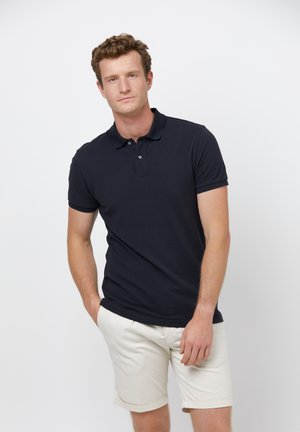Camisa polo de color azul marino con cuello clásico, mangas cortas y tapeta de dos botones; combinada con pantalones cortos de color claro. Diseño simple, ajuste casual.