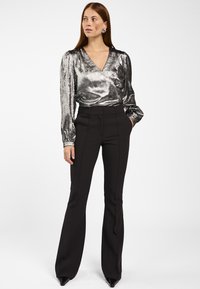 Vrouw met lang rood haar, gekleed in een zilveren metallic blouse met lange mouwen, zwarte hoge taille flare broeken en zwarte puntige schoenen, staand.