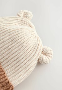 Knit hat in cream with a textured ribbed pattern, features two round ear-like accents on top, and a darker brown ribbed band at the base.
