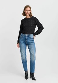 Haut noir à manches longues avec motif texturé ; jean taille haute bleu ciel ; bottines noires à talons pointus. Style minimaliste, silhouette ajustée.