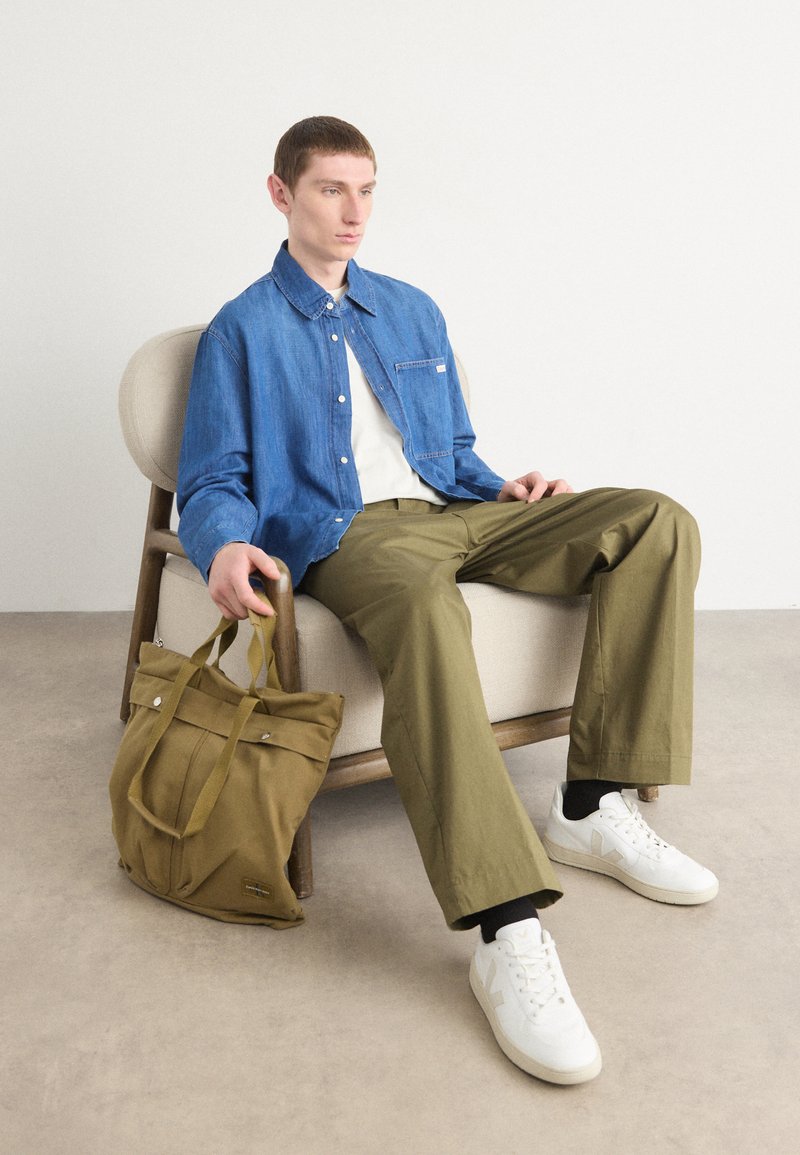 Denim Hemd mit Knöpfen, weißes T-Shirt, olivgrüne Hose, weiße Sneaker. Stofftasche mit zwei Tragen, Details an der Vorderseite und Logo.