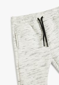 Pantalones de chándal gris claro con un patrón moteado negro, cinturilla elástica y cierres de cordón. Cuenta con bolsillos laterales y una textura suave.