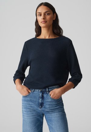 Dunkelblaues strukturiertes Strickpullover mit weitem Ausschnitt und Dreiviertelärmeln, kombiniert mit hellblauen Jeans. Hände in den Taschen.