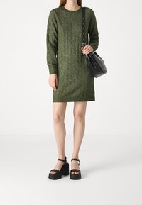 Abito lavorato a maglia verde scuro con un motivo a diamante testurizzato, maniche lunghe, scollatura rotonda, abbinato a sandali con zeppa neri e una borsa nera.