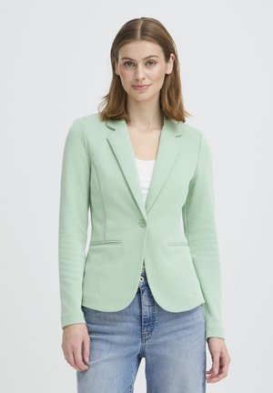 Kobieta w jasnopistacjowej dopasowanej marynarce, białej bluzce i niebieskich dżinsach, stojąca na tle jednolitego jasnoszarego tła.