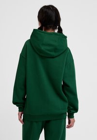 Zielona bluza z kapturem wykonana z miękkiego materiału, z dużym kapturem, luźnym krojem i ściągaczami przy rękawach. Widok z tyłu pokazuje minimalne szwy i brak widocznych znaków marki.