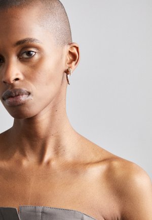 Sidabrinės auskarai-žiedai su kreiviu siluetu, puikuojantys kampuotais akcentais ir subtiliu blizgesiu, dėvimi ant modelio minimalistinėje fone.