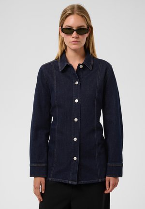 Femme portant une veste en jean bleu foncé boutonnée et des lunettes de soleil noires, se tenant devant un fond clair uni.