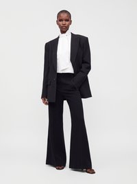 Personne portant un blazer noir oversize, un pantalon noir à jambes larges, une blouse blanche à col montant avec un nœud à l'avant et des talons ouverts noirs.