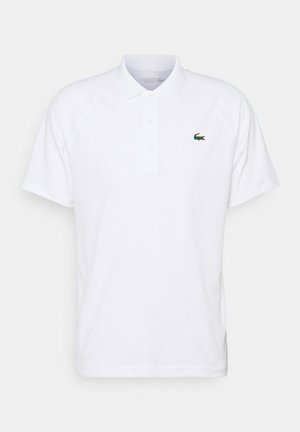 Hvid polo shirt med klassisk krave, tre-knap lukning og grønt Lacoste krokodillelogo på brystet. Korte raglanærmer.