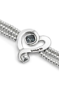 Pulsera de plata con un cierre en forma de corazón que presenta una piedra preciosa gris facetada, rodeada de curvas pulidas y un diseño de cadena con cuentas.