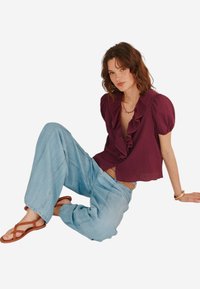 Blusa borgogna con rouches e maniche corte a palloncino abbinata a jeans a gamba larga di colore blu chiaro e sandali marroni con cinturini. Tessuto di consistenza liscia.