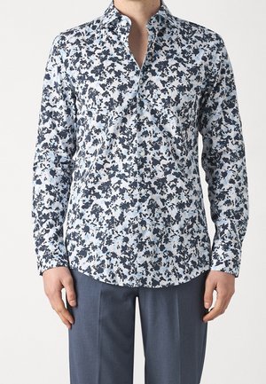 Muž v bílé košili s knoflíky s úzkým střihem a tmavě modrým květinovým vzorem a šedých kalhotách stojící na jednoduchém pozadí.