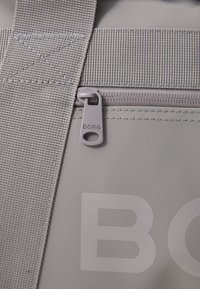 Saco de tecido cinza com uma alça larga, apresentando um fecho em zip e um puxador de zíper marcado "BORG." Superfície texturizada e detalhes de costura duráveis.