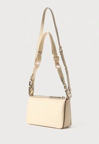 Geantă de umăr din piele bej, cu formă rectangulară, curea reglabilă, închidere cu fermoar și accesorii metalice. Textură netedă.