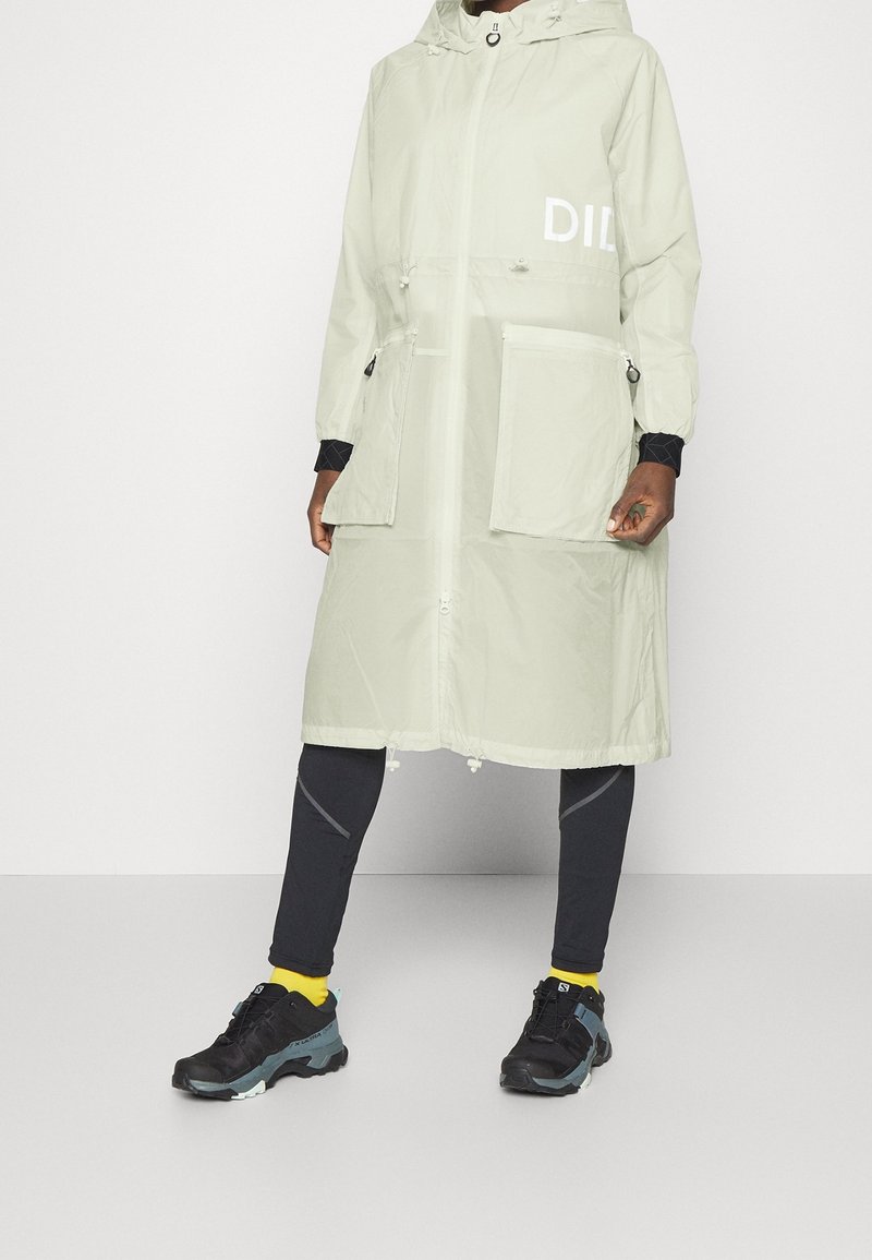 Personne portant un long manteau à capuche beige clair avec de grandes poches avant, des leggings noirs, des chaussettes jaunes et des chaussures de randonnée noires.