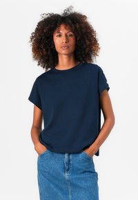 Marineblå topp med korte ermer i myk stoff, med rund hals og gullknapper på skuldrene, sammen med et jeansskjørt.