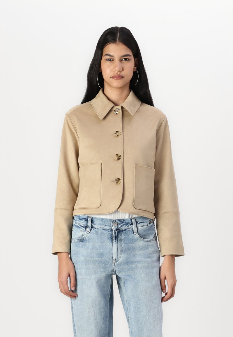 Veste beige courte en tissu lisse, avec un col, cinq boutons et deux poches avant. Associée à un jean bleu clair.