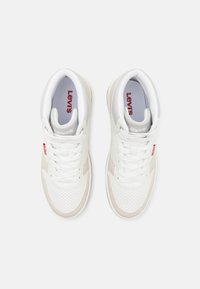 Sneaker in pelle bianca con punta forata, accenti beige e lacci bianchi. Presenta il logo rosso "Levi's" sul lato e sulla soletta.