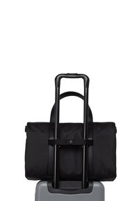 Crna putna torba od tkanine s dualnim ručkama odlaže se na sivi kofer. Ima glatku teksturu i sigurnosnu traku. Vidljiva teleskopska ručka.