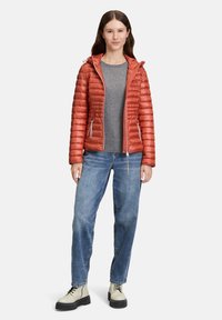 Rød pufferjakke med vandret syninger, lynlås foran, sidelommer og hætte, sammen med en grå skjorte, blå jeans og hvide støvler.