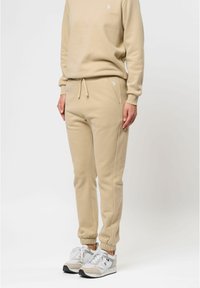 Beige sweatshirtbyxor med elastiska muddar och dragsko i midjan, kombinerade med vita sneakers med grå accenter och nätpaneler.